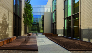 A modern courtyard features glass walls, wooden benches, a tree, and a minimalist design, creating a tranquil outdoor space.