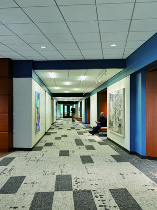 A modern hallway with polished wood and blue accents features abstract artwork, carpeted flooring, and a few people sitting and walking.