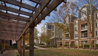 A walkway framed by wooden structures leads to apartment buildings surrounded by trees and greenery under a clear sky.