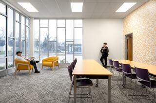 A bright, modern room features two seating areas with yellow chairs, a long table, and a person leaning against the wall, creating a welcoming atmosphere.