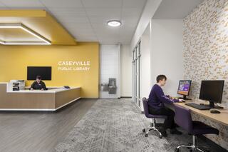 A modern library interior features a reception area with a desk and a visitor using a computer, accented by warm colors and contemporary design.