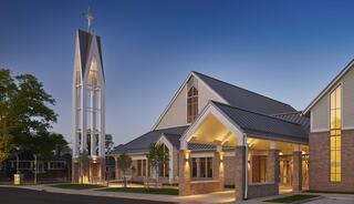 A modern church building features a tall steeple, large windows, and elegant lighting, set in a serene outdoor environment.