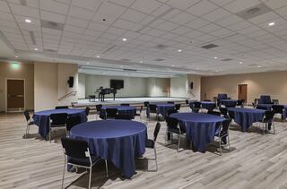 A spacious event room with multiple round tables, dark blue tablecloths, and a stage featuring a piano in the background.