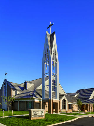 A modern church building features a striking tall spire with a cross, surrounded by green grass and a clear blue sky.