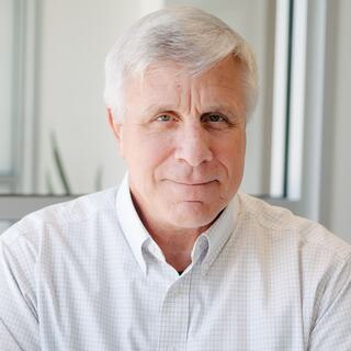 A man with gray hair and a friendly expression wears a light-colored shirt, seated indoors with soft lighting and greenery in the background.