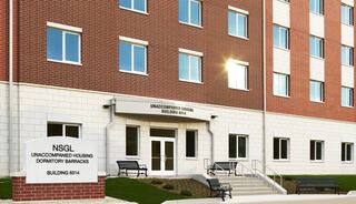 A modern multi-story building features large windows, a welcoming entrance, and clear signage for unaccompanied housing. Benches are placed outside.