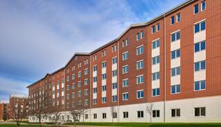 A large, brick building features a symmetrical design with multiple windows and a grassy area in front, set against a cloudy sky.