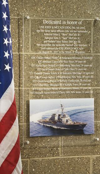 A plaque honors USS John S. McCain and its crew, listing names of individuals recognized for their service and dedication. A flag is present.