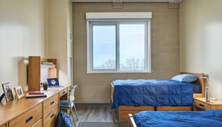 A cozy dorm room with two twin beds, wooden furniture, and a large window offering a view of trees, creating a welcoming atmosphere.