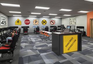 A modern classroom features traffic signs on the walls, with computer stations and bright orange chairs for interactive learning about road safety.