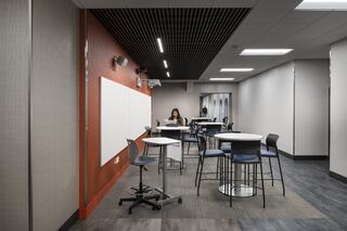 A modern learning space features high tables and chairs, a whiteboard, and a solitary student focused on work. Bright, inviting atmosphere.