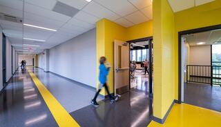 A modern hallway features yellow accents, with students visible in a classroom. A person walks past open doors in a bright, spacious setting.