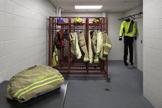 A utility room features a metal rack with fire gear, helmets, and a bright yellow suit, set against plain white walls and a gray floor.