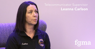 A woman with dark hair sits in a chair, wearing a jacket with a badge, discussing her role as a Telecommunicator Supervisor.