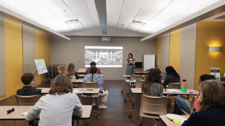 A group of people sits in a seminar room, attentively listening to a speaker presenting on a large screen.