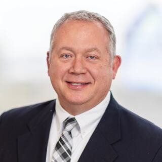 A man in a suit and tie smiles confidently, showcasing a professional appearance with a neutral background.