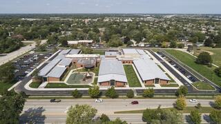 An aerial view of a modern school building surrounded by greenery, parking lots, and nearby residential areas under a clear blue sky.