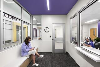 A modern waiting area features a woman sitting on a bench, checking her phone, while another person is seen at a reception desk.