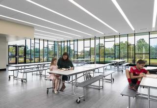 A bright, modern lunchroom features two children reading at a table while a third child sits nearby, enjoying a book in a spacious setting.