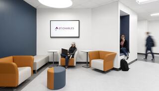 A modern lounge with orange and gray seating, a large screen displaying "St. Charles," and students engaged in various activities.
