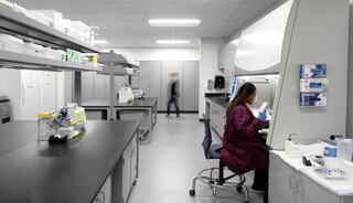 A modern lab with a researcher in a maroon lab coat working at a bench, surrounded by shelves of equipment and supplies. A person walks in the background.