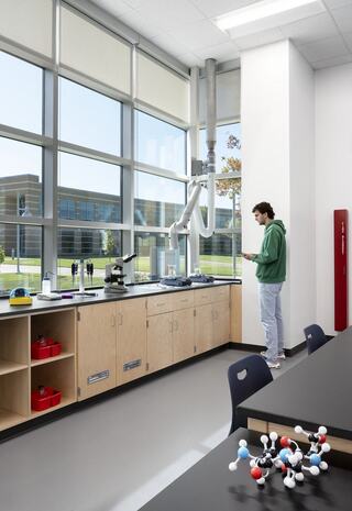 A student stands in a bright laboratory, engaging with a mobile device while a microscope and equipment are nearby, with large windows offering a view outside.