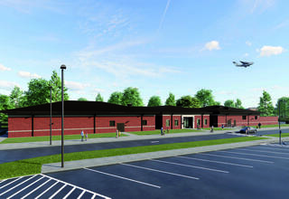 A modern red-brick building with large windows, surrounded by green trees, parked cars, and an airplane flying overhead.