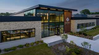 A modern building features large glass windows and a prominent wooden sign, surrounded by landscaped greenery. It's inviting and contemporary.