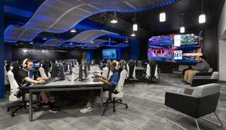 A modern esports gaming arena features several players engaged at computer stations, with a large screen showcasing a tournament in the background.
