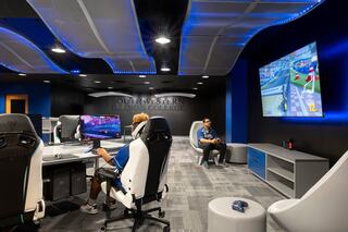 A modern esports gaming arena at Southwestern Illinois College features sleek chairs and multiple screens, with students engaged in gaming and relaxed on stylish seating. One student plays a computer game and another plays on a console with a large TV screen.