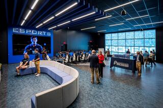 A modern college esports arena features gaming stations, a curved sofa for console gaming, and a large screen showcasing players, with attendees mingling and discussing.