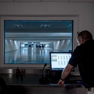A person monitors a shooting range through glass, observing three individuals in uniform practicing on the range, with computer data displayed nearby.