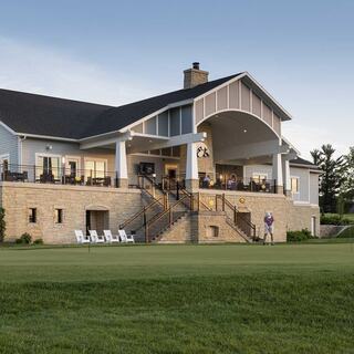 A charming golf clubhouse features a spacious porch, inviting seating, and lush green surroundings, perfect for relaxation and socializing.