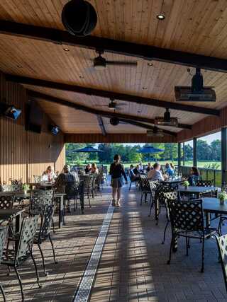 A bustling outdoor dining area features elegant tables and chairs, with guests enjoying the ambiance under a wooden ceiling and ceiling fans.