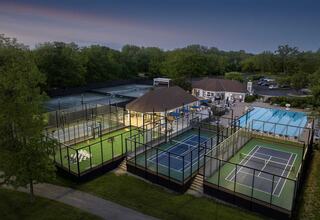 A sports complex featuring multiple courts, including tennis and padel, surrounded by greenery and a central clubhouse, illuminated at dusk.