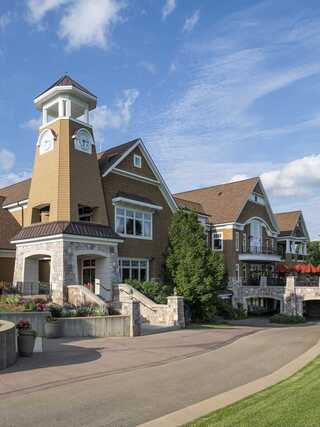 A charming, multi-story building features a clock tower and stone accents, surrounded by lush greenery and a scenic pathway.