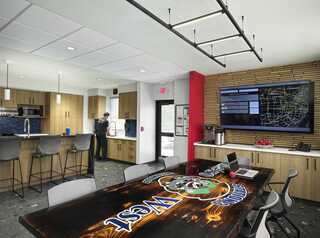 A modern kitchen and dining area at the renovated Metro West Fire Station 3, featuring a large branded dining table, sleek cabinetry, and a TV displaying a map of the Metro St. Louis, Missouri area..