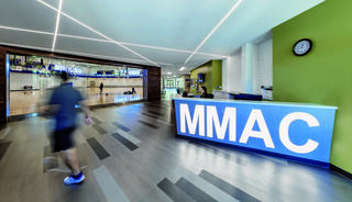 A bright entry lobby at Madison Meadows Athletic Center features a large "MMAC" sign at the reception desk, sleek flooring, and a glimpse of an active gym area in the background.