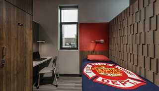 A modern, compact fire station bunk room featuring wooden accents, a desk, and a single bed with a fire department-themed blanket against a textured wall.