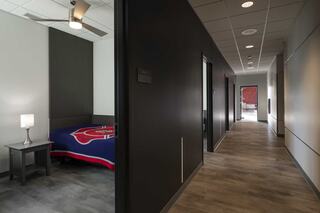 A sleek, modern hallway features doorways to fire station bunk rooms, with a bedroom visible showing a bedside lamp and a colorful blanket on a single bed.