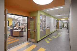 A bright school hallway leads to a colorful library and media center with playful decor, welcoming seating areas, and vibrant wall art. The hallway is painted in yellow, green and orange, and green circles make a dotted path to the media center, with orange rectangles leading into the center and beyond.