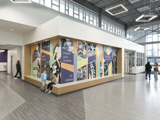 Bright window-filled lobby with a corner mural and a corner seating bench at Caseyville Elementary School. Students and faculty are coming and going, and two young students sit on the corner bench. The mural features orange, purple, yellow and light teal colors, with welcoming and inviting phrases in both English and Spanish, as well as black and white photos of the local community.