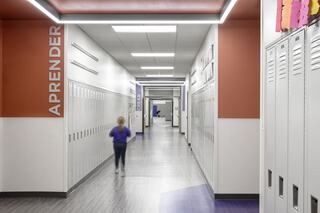 Caseyville Elementary School hallway lined with tall lockers on each side featuring a student walking away. Walls have orange or purple paint depending on the space type associated with the color and bilingual English and Spanish signage is seen throughout the hallway.