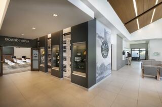 Village of Cary, Illinois' new village hall and police combined facility lobby, showing two paths in the bright lobby with a sign labeled "Board Room" above an open door and a person standing at an administrative desk. Glass cases line one wall, showing off historical items. A custom graphic of historic images lines the wall behind the Village's seal.