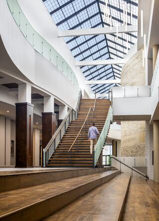 A person ascends a wooden staircase in a modern, spacious interior with large windows and elegant design elements.