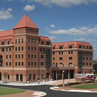 A modern brick building with a red tiled roof features multiple stories, large windows, and an accessible entrance, set against a blue sky.