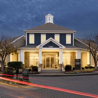 A charming two-story building with a peaked roof, warmly lit windows, and landscaped entry, featuring a leasing office sign out front.