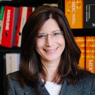 A woman with long, dark hair and glasses smiles warmly, wearing a blazer, in front of shelves filled with colorful folders.