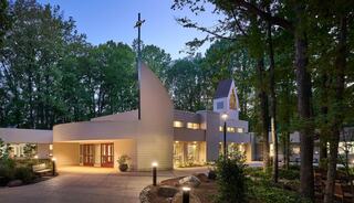 A modern church with a cross atop, nestled among trees, features warm lighting and a welcoming entrance, creating a serene atmosphere.