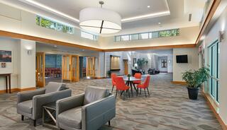 A bright, modern multi-purpose gathering space featuring comfortable seating, a stone fireplace, large windows, and lush greenery outside, creating a welcoming atmosphere.